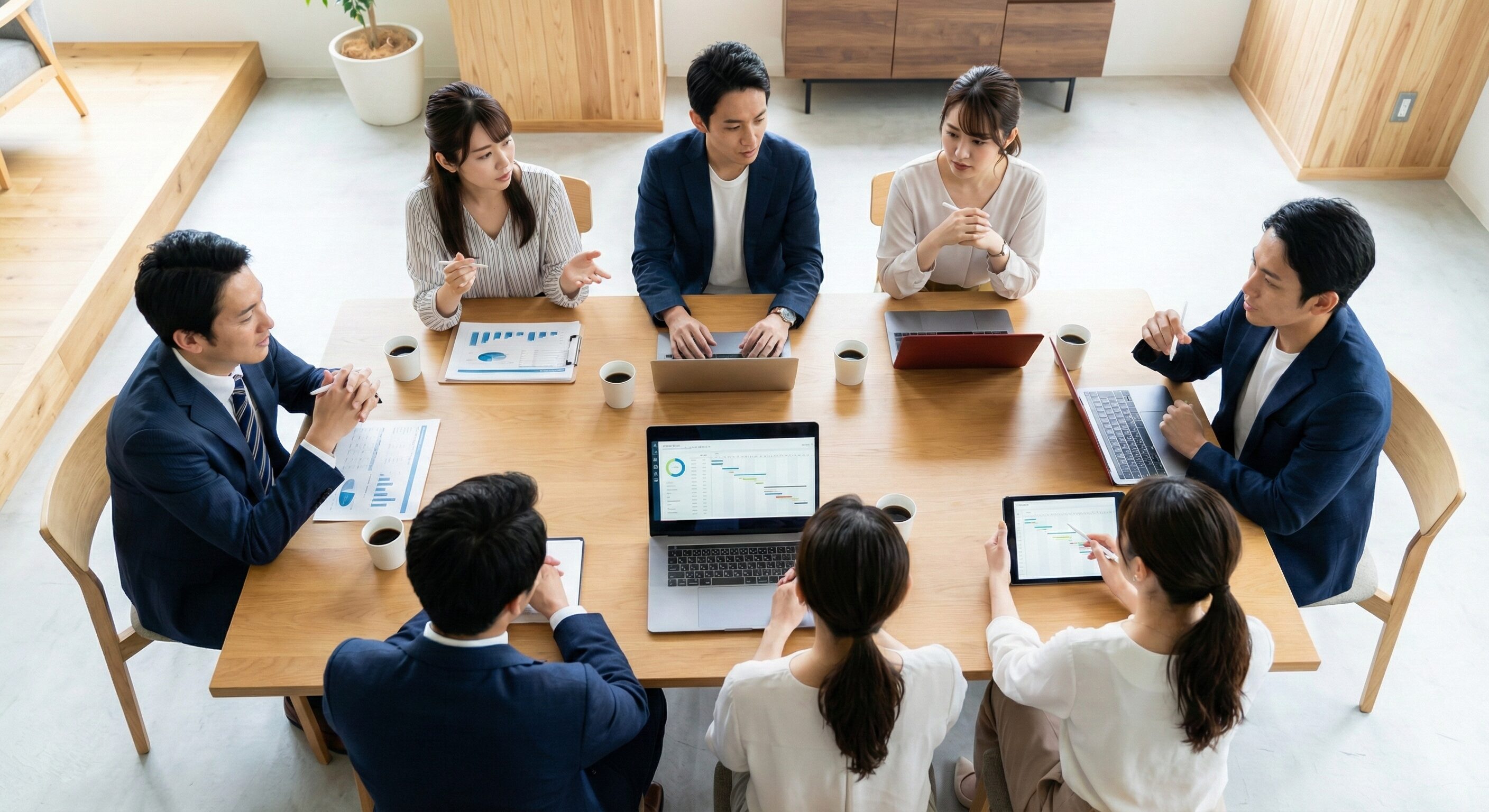 複数のビジネスパーソンが会議テーブルを囲み、ノートPCやタブレットを使いながら真剣に議論している俯瞰写真。チームワークと目標達成への意志を感じる構図。